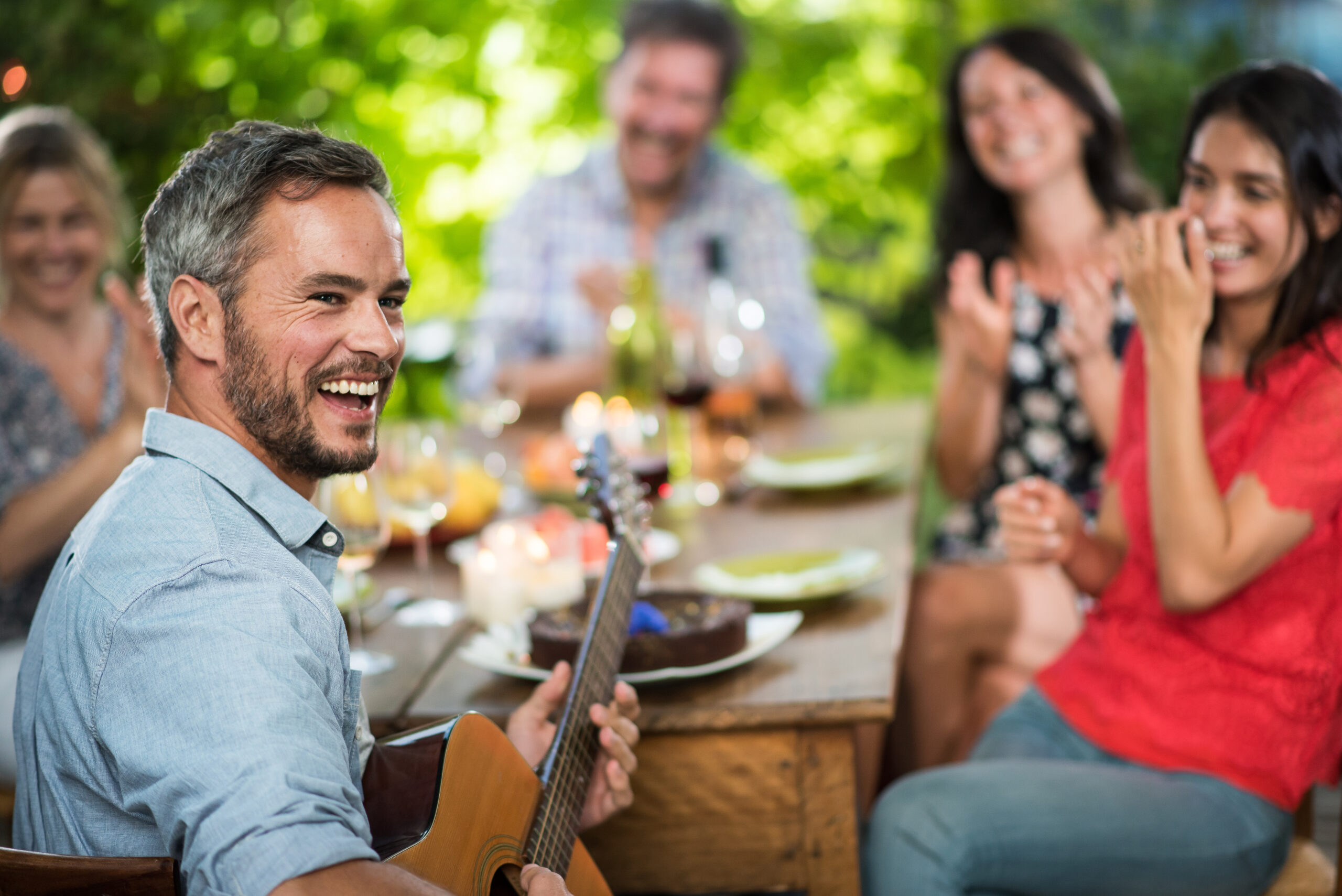 Summertime. Handsome gray haired man enjoying playing guitar for his friends. They gathered around a terrace table where they shared a meal