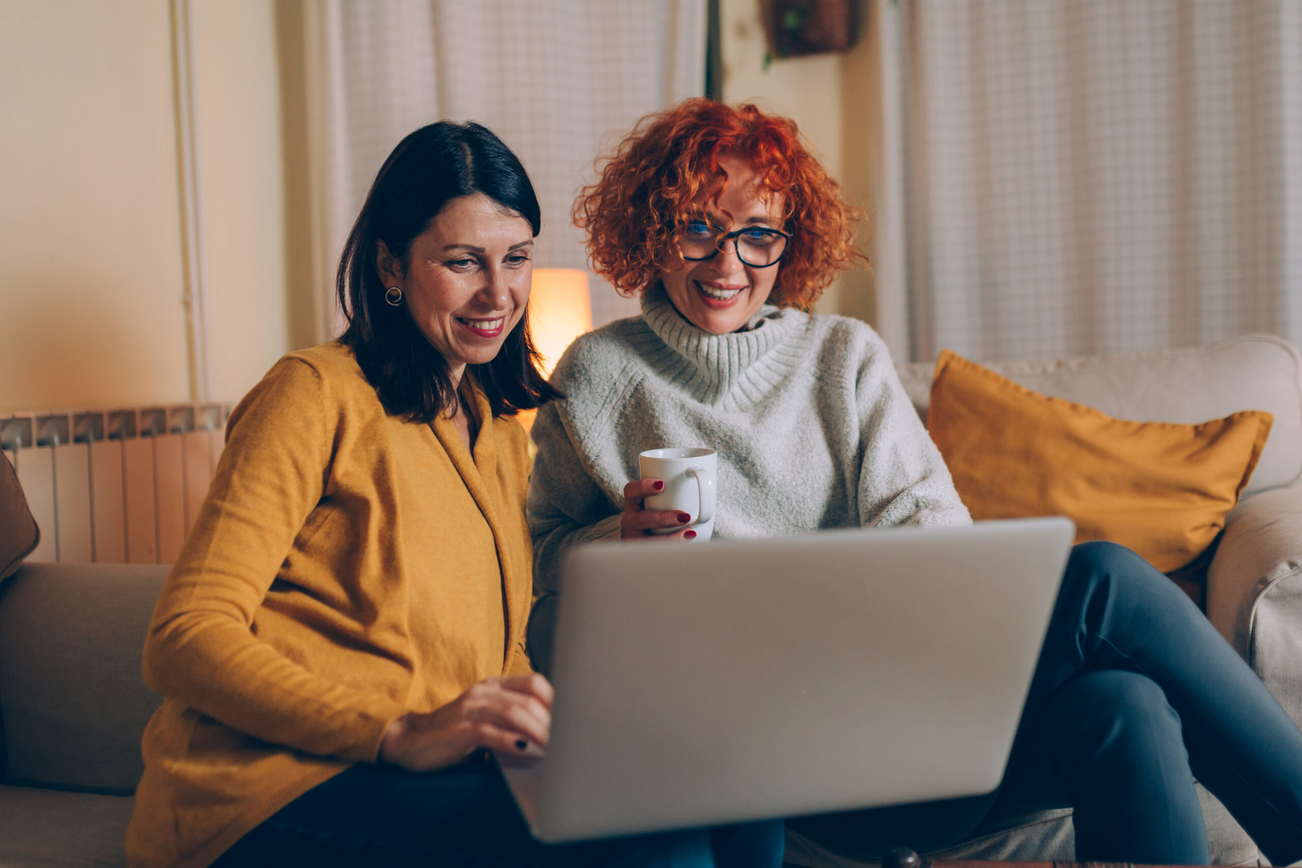 woman friends using laptop at home, sitting sofa drinking coffee and planning